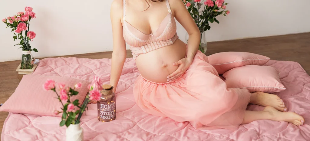 Mulher grávida, sentada de lado em lençol rosa no chão em cenário romântico - Just Real Moms