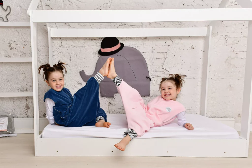 Meninas sorrindo deitadas em cama infantil usando saco de dormir azul marinho e outro rosa