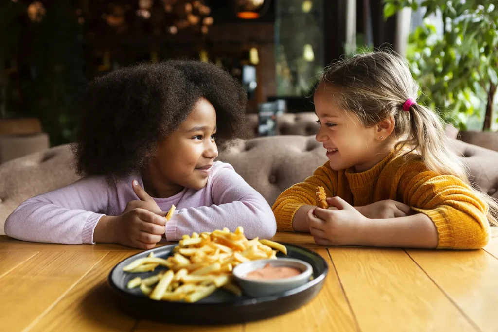 Duas meninas amigas sorrindo e sentadas a mesa comendo batata frita nas férias