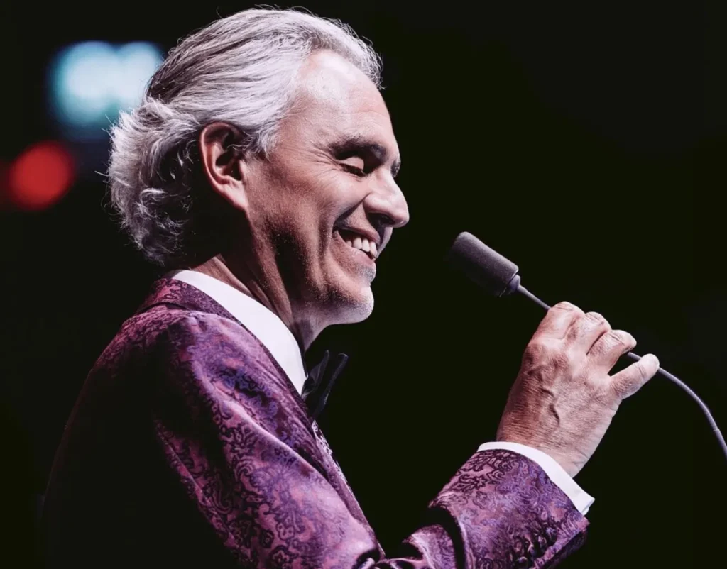 Close de Andrea Bocelli no palco sorrindo com microfone na mão usando blaser lilás