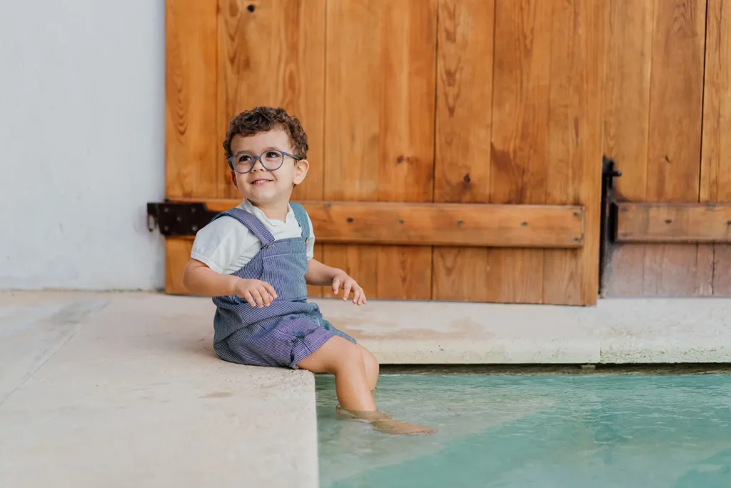 Menino bebê de óculos, sorrindo com pés dentro de uma piscina interna, vestindo Little Jam