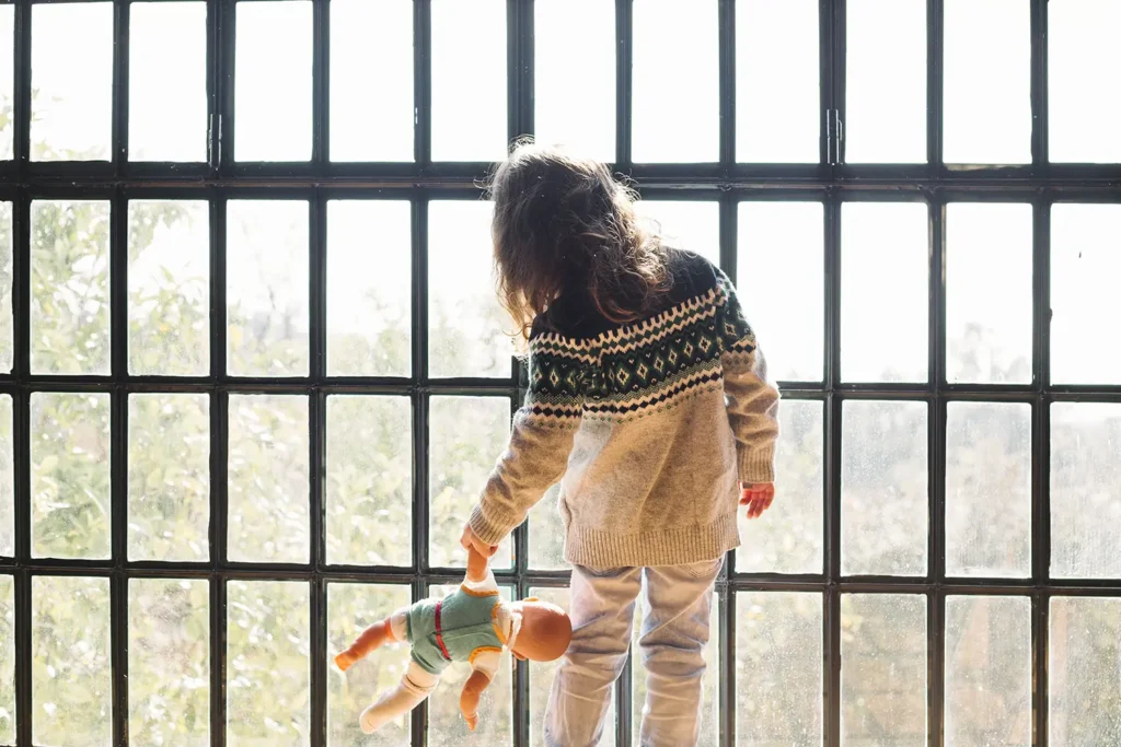 Menina olhando para fora de uma janela, segurando uma boneca - Alergia alimentar em crianças