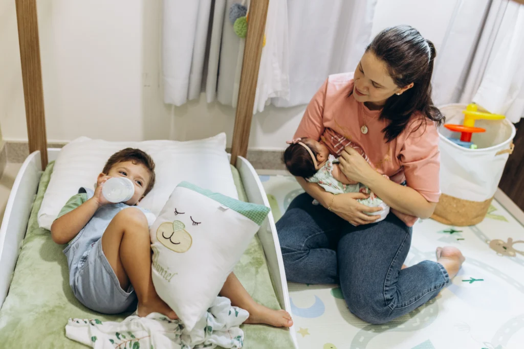 Mãe de dois filhos amamentando o seu bebê enquanto põe o mais velho para dormir em um quarto cercado de produtos para gestantes