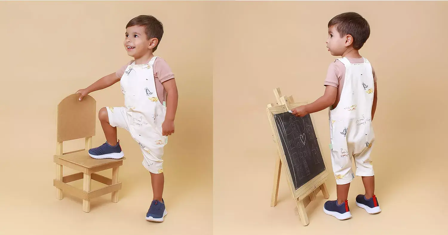 Menino de macacão ao lado de cadeira e ao lado de quadro de giz, vestindo Mimor